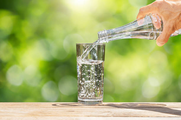 hand holding drinking water bottle pouring into glass on wooden table on blurred green nature background - water water bottle glass pouring imagens e fotografias de stock