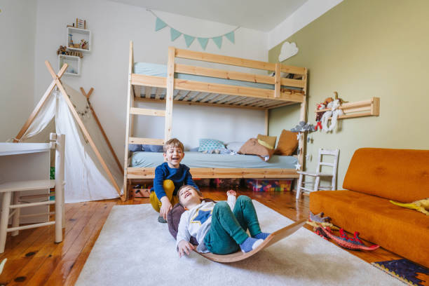 Two little boys playing in their room Photo of two little boys playing with balance board in his room kids room. Colorful and cozy kids room featuring a white bunk bed with blue and green bedding, a red and white striped rug, a wooden toy chest, a bookshelf filled with children's books, and playful wall decor including a colorful animal print and a growth chart