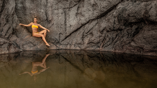 A woman in a yellow bikini poses on the rock pool Ros Sodyer on Mahé Island. As it is early morning the water is very calm and the model is reflected on the surface. During every high tide the pool's water is renewed.