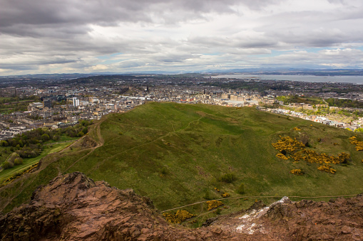 View from Arthurs Seat looking over Holyrood Park with the Edinburgh city center and the Firth of Forth in the background, lightened by dappled light of sun shining through a cloudy sky