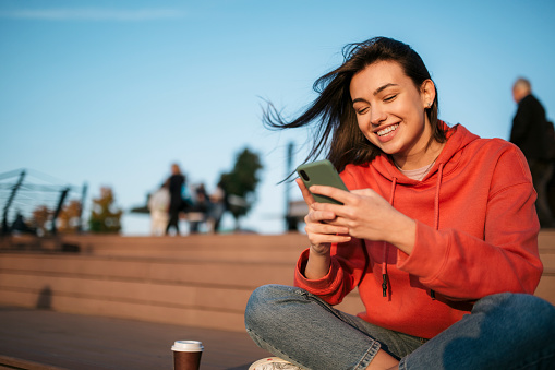 Happy young Caucasian woman relaxing outdoors in a red shirt, smiling and using a smart phone