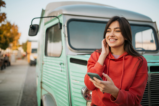 Happy young Caucasian woman standing outdoors, in front of an old truck and in a red shirt, smiling and using a smart phone