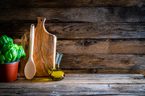 Cooking and kitchen backgrounds: front view of a wooden cutting board, basil, olive oil and a spoon on rustic wooden table. The composition is at the left of an horizontal frame leaving useful copy space for text and/or logo at the right. High resolution 42Mp studio digital capture taken with SONY A7rII and Zeiss Batis 40mm F2.0 CF lens