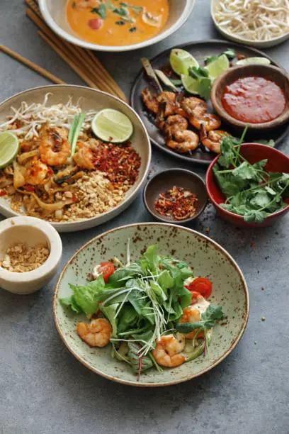 Spicy shrimp salad. Authentic classic Pad Thai with shrimps. Thai shrimp satay with sweet chili sauce. Tom Yum soup with coconut milk (Tom Khaa). Close-up composition on concrete background.