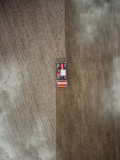 縦の垂直赤いトラクターは、雲を通して上から見下ろす農場のフィールドの空中写真を耕します。農家の春の植栽によって駆動される田舎の耕作農地の耕作作業ドローン写真栽培機機器 - tractor farm uk agriculture ストックフォトと画像