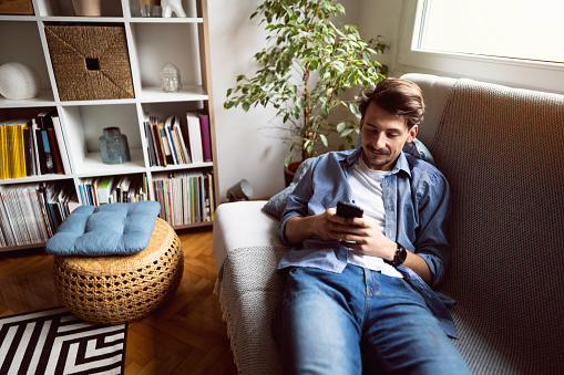 One handsome young Caucasian man lying on the sofa at home and using a smart phone.