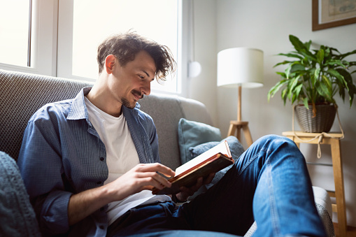 One handsome young Caucasian man sitting on the sofa at home and reading a book.