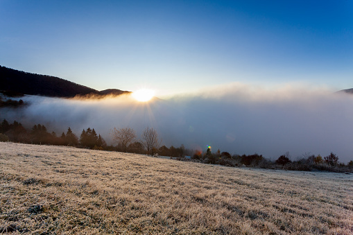 Sun rises from behind mountain top, fog moves and rises up at dawn on summer morning in mountains. Morning fog dissipates in mountains Carpathians. Nature landscape, natural scenery, rural background.