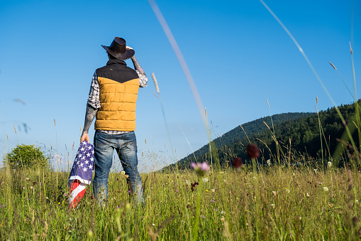 Rear view on cowboy with sleeve tattoos holding his hat and American flag looking at the field in front.