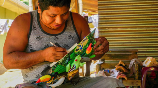Local artist from the Boruca tribe giving a signature on his artwork. Puntarenas Province, Costa Rica-March 2018: Local artist from the Boruca tribe giving a signature on his artwork piece to a tourist who is buying it. Traditional mask made from wood is a famous crafting souvenir that these people are making. traditional sun symbol art for sale stock pictures, royalty-free photos & images