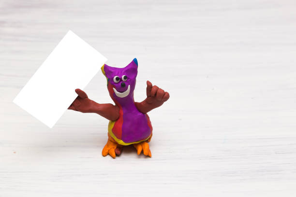 istock A plasticine figure resembling a cat on a light background holds a clean empty table. Blank sheet. Copy space 1310634393