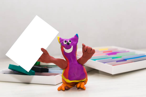 istock A plasticine figure resembling a cat on a light background holds a clean empty table. Blank sheet. Copy space 1310634440