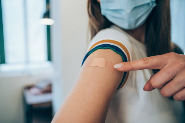 Woman showing her arm after getting vaccinated. Woman wearing protective face mask and pointing at her arm with a bandage after receiving COVID-19 vaccine. Young woman showing her shoulder after getting coronavirus vaccine at vaccination center. adhesive bandage stock pictures, royalty-free photos & images