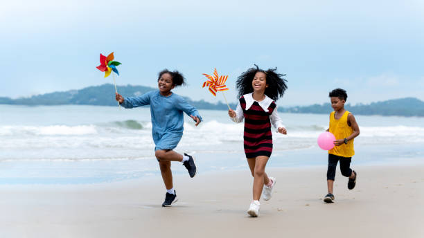 feliz niño afroamericano corriendo con rueda de alfiler y disfrutando con una pelota de playa en la playa tropical - run of the mill fotografías e imágenes de stock