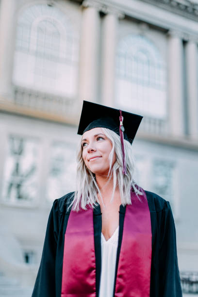 Low Angle Portrait of a Confident College University Graduate with Copy Space Portrait of a Happy, Confident, Successful College University Graduate Young Woman Wearing a Maroon School Color Sash and a Mortarboard Graduation Cap and Tassel at Sunset Outdoors golden hour wine stock pictures, royalty-free photos & images