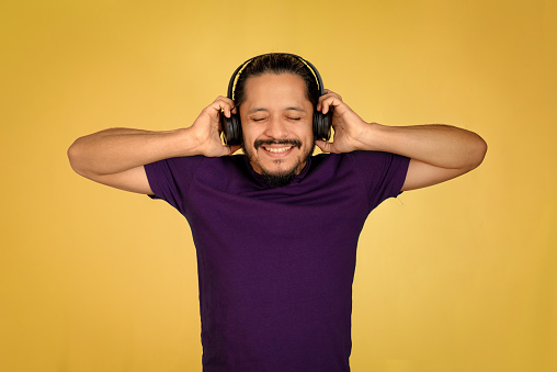 Young man using headphones smiling with his hands on the ears with yellow background