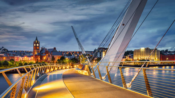 Northern Ireland Derry The Peace Bridge River Foyle Night Panorama Londonderry Illuminated Londonderry 'The Peace Bridge' over the River Foyle in Derry (Londonderry ) at Twilight. Panorama of the modern architecture Peace Bridge in Derry over the River Foyle after during overcast twilight with Guildhall and Old Town Cityscape in the background. The Peace Bridge is a cycle and foot bridge across the River Foyle in Derry - also called Londonderry, Northern Ireland, Europe northern ireland photos stock pictures, royalty-free photos & images