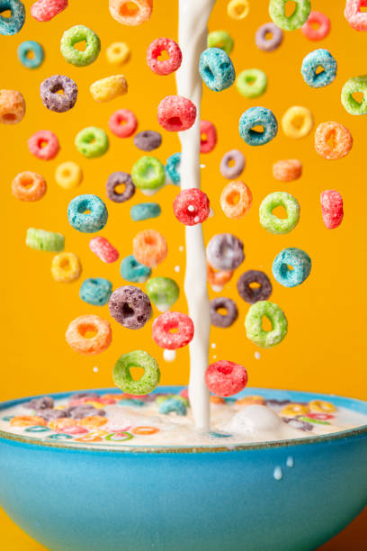 Milk and cereal poured in a bowl. Preparing breakfast, creative layout Dropping cereals and pouring milk into a blue bowl, isolated on an orange background. Preparing breakfast with flying colorful cereals and milk drip. vibrant color lifestyles vertical close up stock pictures, royalty-free photos & images