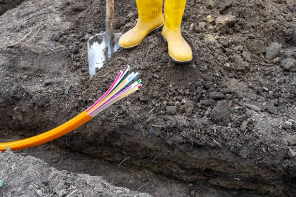 Construction worker with yellow rubber boots stands in front of a trench in which an orange plastic cable for optical fiber is lying