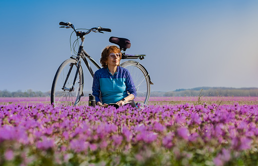 Older woman bicyclist in colorful jersey enjoying nature while sitting in field of purple wildflowers; bicycle behind her