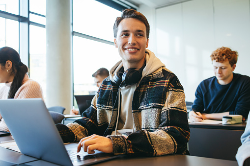 Young man sitting at classroom desk looking away and smiling. Student with laptop on desk in class.