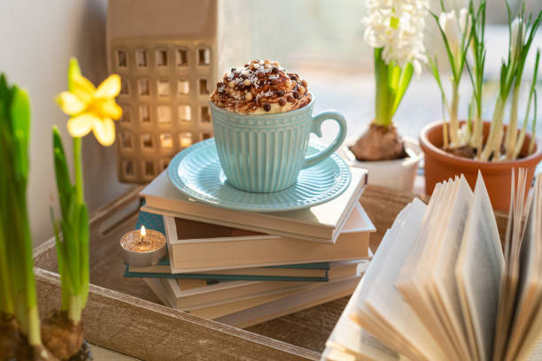 Coffee cup with whipped cream, spring flowers and open book on window sill. Concept warm and cozy home, hygge, reading books, rest and comfort, springtime