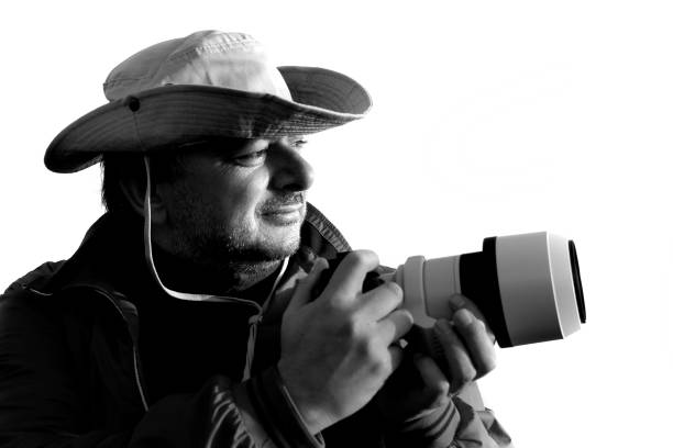 A photographer holds his camera and looking away. He is isolated whit white background. He wears a hat.