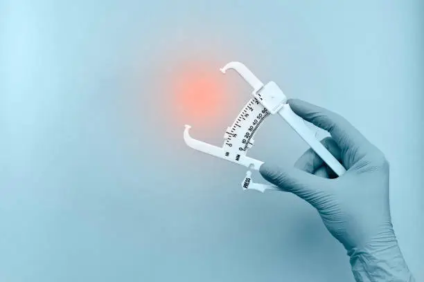 Caliper for measuring the thickness of subcutaneous fat in the hand, with a rubber glove, in light blue tones. With a red glowing accent at the research site.