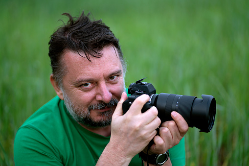 A man is holding his camera and looking at the objective. He is wearing green shirt in front of green grass background.