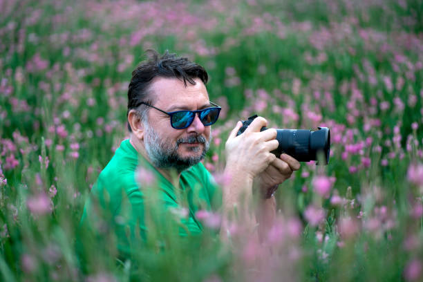A man wearing sunglasses and green shirt. He is sitting in the pink flowers garden. He is holding his camera.
