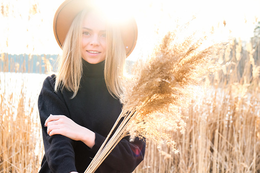 European blonde woman with beige hat in black sweater in the countryside. Golden hour, cottagecore. Local travel. Slow living. Mental health. Earth tones. Pampas grass