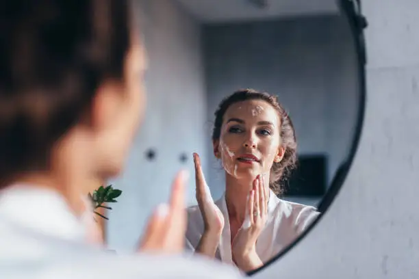 Woman washes in front of the mirror, applying foam to her face.