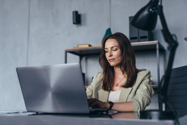 mulher trabalha em seu escritório sentado em sua mesa com um laptop - women business serious businesswoman - fotografias e filmes do acervo