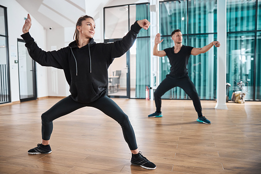 Sportswoman and male both doing tai chi single whip exercise while standing on wooden floor of vacant office room