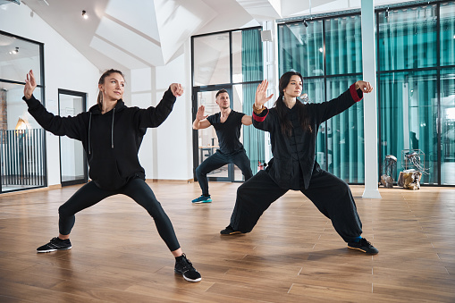 Female tai chi practitioner giving example of single whip stance to man and woman following her instructions