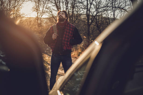 un hombre barbudo camina por el bosque mientras la puesta de sol está detrás de él. un hombre, un leñador, va a su cabaña de troncos, que está en un bosque profundo. - lumberjack shirt fotografías e imágenes de stock