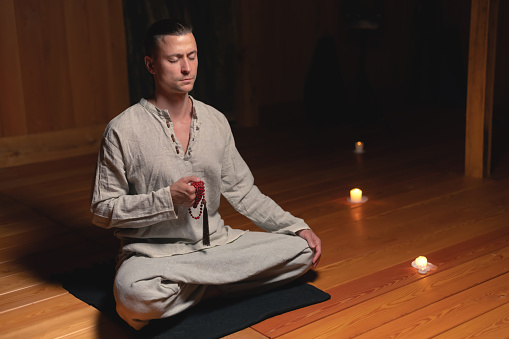 A young attractive man in gray natural clothes sits in a lotus position in a dark wooden practice room with a red rosary in his hand. Eyes closed practice and religion.