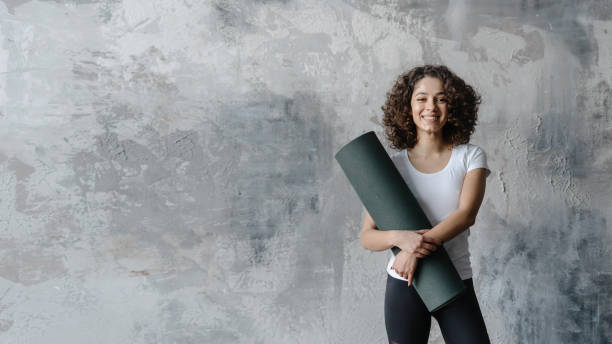 Young african american woman standing with exercise mat in fitness center Confident and professional coach in sportswear standing with rolled exercise mat against copy space background. Happy young african american woman preparing for workout training in sport club teenage yoga stock pictures, royalty-free photos & images