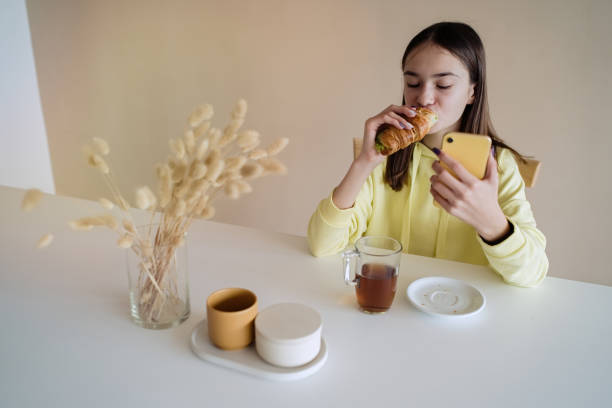 Teen girl eating breakfast in kitchen while using on her smart phone