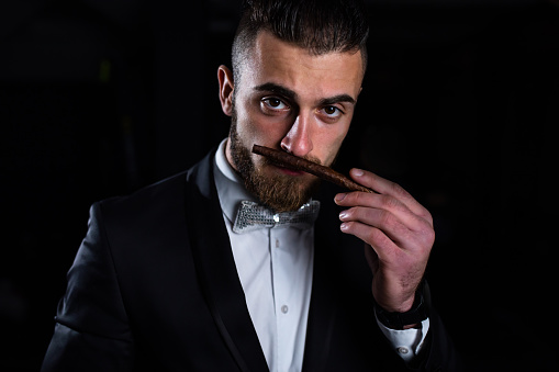 A handsome man in a suit is looking at the camera and smelling a cigar in the dark