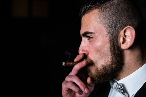 A handsome man in a suit is smoking a cigar in the dark