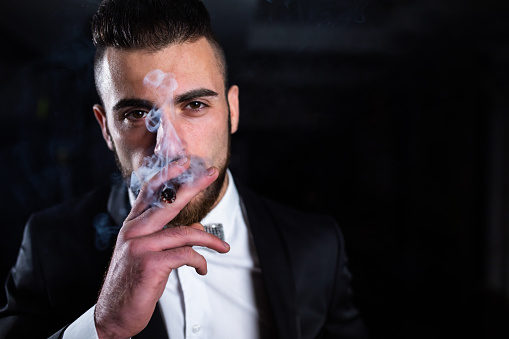 Rude and impudent man scoundrel in formal suit and smoked sunglasses sits on chair backwards smoking cigarette looks down at camera over dark background. Stylish look for men concept