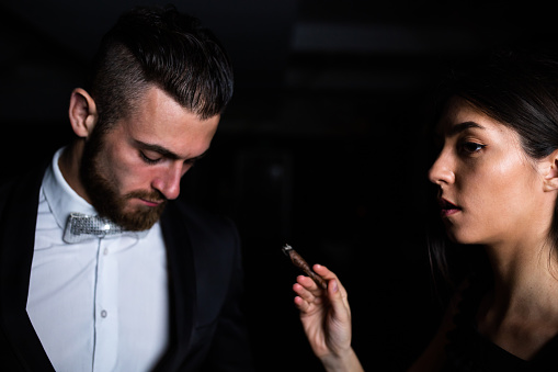 A handsome young man is looking down while a petty young woman is smoking a cigar next to him
