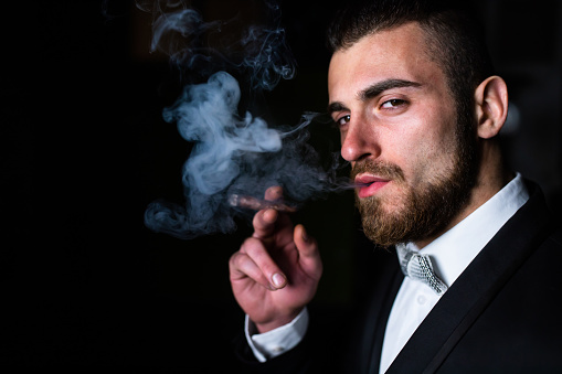 A handsome man in a suit is smoking a cigar in the dark