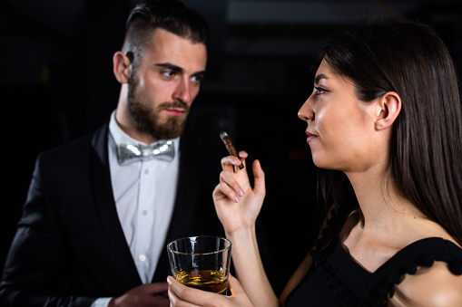 A young woman is holding a glass of scotch and smoking a cigar next to a handsome young man in a suit
