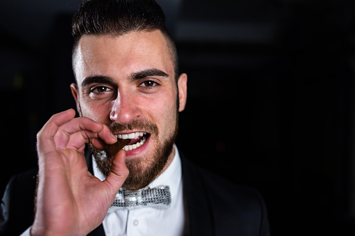 A handsome young man in a suit is holding a cigar between his teeth in the dark