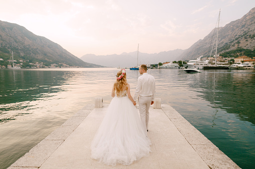 The bride in a wreath and groom walk along the pier holding hands near the old town of Kotor in the Bay of Kotor . High quality photo