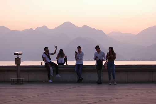 05.11.2019 - Antalya, Turkey. Tourists on the embankment at resort town during sunset. Young man taking selfie on the background of a sea and mountains. Antalya, Turkey