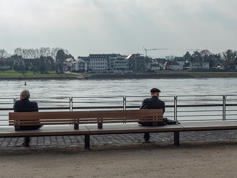 Neuwied, Germany - March 23, 2021: two men seen from behind, sitting with distance on a bench at the quayside of the Rhine - symbol for loneliness and social distancing in times of the Corona pandemic
