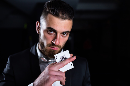 A handsome man in a suit is holding the ace of spades between his fingers in the dark